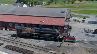 Strasburg, Pennsylvania, 28 Haziran 2022 - Vintage lokomotif bir demiryolu atölyesinin önünde bakım geçiriyor. Arkaplanda yemyeşil alanlar görülebiliyor, cennet gibi bir alanı yakalıyorlar.
