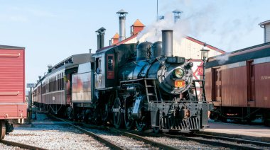 Strasburg, Pennsylvania, 24 Şubat 2024 - Antika bir buhar treni tuhaf bir tren istasyonundan kalkmaya hazırlanıyor. Parlak öğleden sonra güneşi aktif motorları ve yükselen dumanı vurguluyor