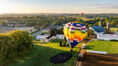 Güneş doğarken huzurlu bir kırsal bölgede canlı bir sıcak hava balonu yükselmeye hazırlanıyor. Çiftlikler ve ağaçlar fırlatma alanının çevresini sararak, güzel bir sabah manzarası yaratıyor..