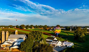 Renkli bir sıcak hava balonu sakin kırsal bir arazide gökyüzüne yükselir. Çiftlik evleri ve tarlalar batan güneşin sıcacık parıltısıyla aydınlanıyor..