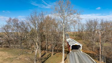 Tarihi bir köprü, çıplak ağaçlar ve renkli sonbahar yapraklarıyla çevrili kırsal bir alanda sessiz bir yol boyunca uzanır. Sahne sakin ve resimsel bir manzarayı yansıtıyor..