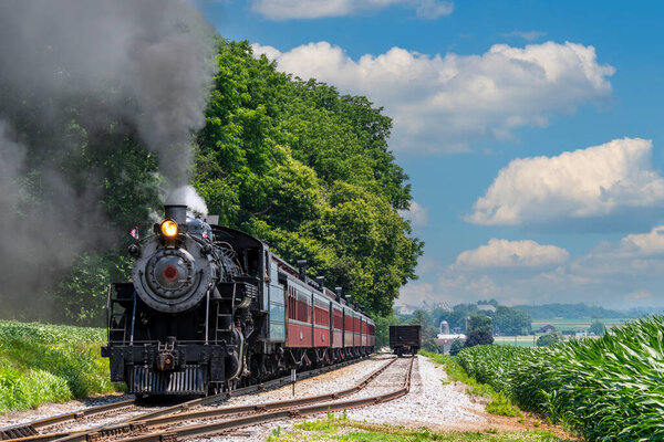 A steam locomotive puffs clouds of smoke as it glides along tracks in a vibrant green landscape. The sun shines brightly and fluffy clouds dot the blue sky, creating a picturesque scene.