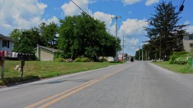 Amish at arabasının yemyeşil mısır tarlalarının yanında kırsal bir yol boyunca ilerlediği görülürken, mavi gökyüzünün altında tüylü beyaz bulutlarla dolu bir sarı araba karşı istikamete yaklaşıyor..