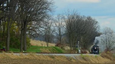 Kırsal bir yolda klasik bir buhar lokomotifi, ağaçların altın ve kahverengi tonlarında yapraklar saçarak kırsal kesimde güzel bir sonbahar günü yaratıyor..