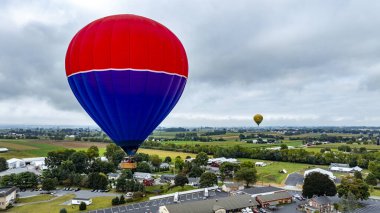 Canlı bir sıcak hava balonu açık arazilerin ve ilginç evlerin üzerinde süzülür. Uzakta görünen başka bir balonla kırsal manzaranın çarpıcı bir görüntüsünü yaratır..