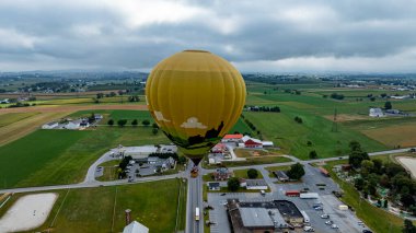 Canlı bir sıcak hava balonu yeşil alanlar ve dağınık binalarla dolu güzel bir kırsal arazide zarifçe süzülür. Bulutlu gökyüzü manzaraya sakin bir atmosfer ekler..