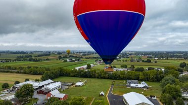 Geniş yeşil alanlar ve bulutlu bir gökyüzünün altında tarım yapıları üzerinde canlı bir sıcak hava balonu süzülür. Kırsal alan, hava manzarası için sakin bir zemin sağlıyor..