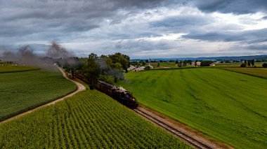 Eski model bir buhar treni uçsuz bucaksız yeşil tarlalar ve bulutlu bir gökyüzünün altında dolambaçlı demiryolu boyunca hareket eder. Manzara, kırsal sükunetin özünü yakalar..