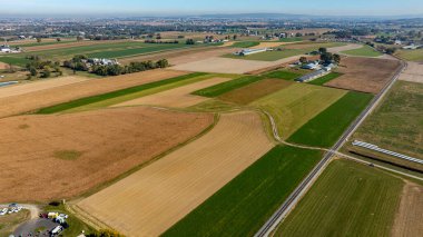 Havadan bakıldığında çeşitli tarlaların farklı renk ve desenler gösterdiği geniş bir tarımsal manzara ortaya çıkıyor. Görüntü, açık mavi gökyüzünün altında kırsal özü yakalıyor..