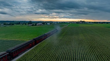 Kırsal bir ortamda, geniş yeşil alanlar arasında bir tren ufukta süzülüyor. Güneş batarken tepemizde fırtına bulutları beliriyor, arazide sıcak bir parıltı yayıyor..