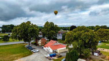Sıcak hava balonu, bereketli ağaçlar ve büyüleyici bir binayla çevrili kırsal bir bölgenin üzerinde zarifçe yükselir. Bulutlu gökyüzü sahneye benzersiz bir ambiyans ekler.