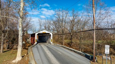 Çıplak ağaçlar ve tarlalarla çevrili sakin bir nehir boyunca uzanan örtülü bir köprü. Gökyüzü açık ve mavidir, kırsaldaki huzurlu sonbahar manzarasını vurgular.