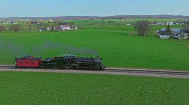 Ronks, Pennsylvania, 1 Nisan 2023, yemyeşil tarlalar ve tarım arazileriyle çevrili raylar boyunca buharlı bir lokomotif tıkınıyor. Bu pitoresk yolculuk doğanın ve tarihin güzelliğini gözler önüne seriyor.