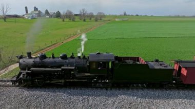 Eski model bir buhar treni, açık bir gökyüzünün altında canlı yeşil tarlalar ve ağaçlarla çevrili raylar boyunca tıkınıyor. Islığı sükuneti deliyor, nostaljiyi çağrıştırıyor.