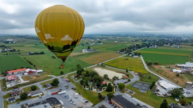 Canlı bir sıcak hava balonu yeşil alanlar ve dağınık binalarla dolu güzel bir kırsal arazide zarifçe süzülür. Bulutlu gökyüzü manzaraya sakin bir atmosfer ekler..