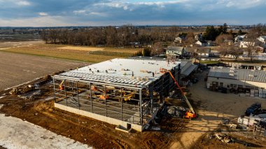 Çalışmalar büyük bir ticari yapı üzerinde devam ederken, işçiler aktif olarak çatı malzemeleri kuruyor ve bulutlu bir gökyüzü altında çelik iskeleti güvence altına alıyorlar. Çevre tarlalar ve evler görülebilir.