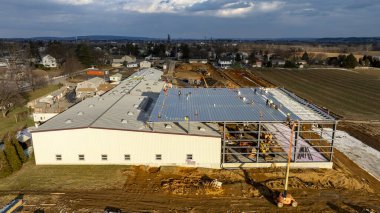 Çalışmalar büyük bir ticari yapı üzerinde devam ederken, işçiler aktif olarak çatı malzemeleri kuruyor ve bulutlu bir gökyüzü altında çelik iskeleti güvence altına alıyorlar. Çevre tarlalar ve evler görülebilir.