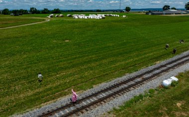 Üç kişilik bir grup tren rayları boyunca yürüyor. Arkaplanda beyaz balyalar bulunan yemyeşil alanlar ile çevrilidir. Gökyüzü bulutlarla açık..