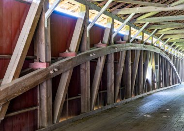 Kapalı bir köprünün iç yapısının çarpıcı bir görüntüsü karmaşık ahşap kirişler ve desteklerle dolu, gün ışığında huzurlu bir ülkede işçiliğin altını çiziyor..