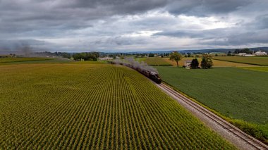Tarihi bir buhar treni, bulutlu bir gökyüzünün altındaki canlı yeşil arazilerden geçerken tren rayları boyunca tıkırdıyor. Kırsal bölgelerde tepeler ve çiftlik evleri sergileniyor..