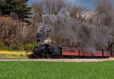 Antika bir buharlı lokomotif, manzaralı bir yol boyunca kırmızı bir yolcu vagonunu çeker. Tren yeşil alan ve bulutlu gökyüzüne duman bulutları yayar ve bir anlığına nostalji yakalar..