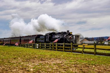 Tarihsel buharlı lokomotif, bulutlu bir gökyüzüne doğru yükselen buhar dumanlarıyla raylar boyunca klasik yolcu vagonlarını çeker. Sahnede yeşil bir tarla ve yakınlardaki tahta çitler yer alıyor..