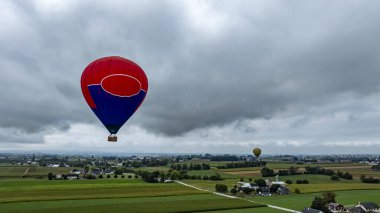 Canlı bir sıcak hava balonu açık arazilerin ve ilginç evlerin üzerinde süzülür. Uzakta görünen başka bir balonla kırsal manzaranın çarpıcı bir görüntüsünü yaratır..