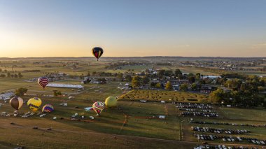Renkli sıcak hava balonları yeşil tarlalar ve park alanlarıyla çevrili bir festival sahasının üzerinden akşam gökyüzüne yükselirler ve kırsal kesimde canlı bir kutlamanın ruhunu yakalarlar..
