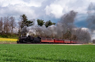 Antika bir buharlı lokomotif, manzaralı bir yol boyunca kırmızı bir yolcu vagonunu çeker. Tren yeşil alan ve bulutlu gökyüzüne duman bulutları yayar ve bir anlığına nostalji yakalar..