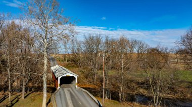 Çıplak ağaçlar ve tarlalarla çevrili sakin bir nehir boyunca uzanan örtülü bir köprü. Gökyüzü açık ve mavidir, kırsaldaki huzurlu sonbahar manzarasını vurgular..