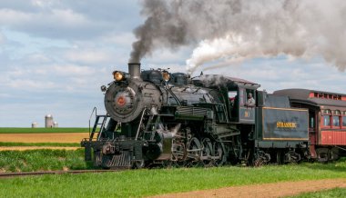 Ronks, Pennsylvania, 13 Nisan 2025 - Eski bir buhar lokomotifi bulutlu bir gökyüzü altında dumanı dalgalandırarak kırsal manzaranın ve tarlaların güzelliğini gün ışığında sergiliyor..