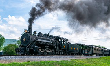 Rockhill Furnace, Pennsylvania, 5 Ağustos 2023 eski bir buhar lokomotifi raylar boyunca seyahat ederek havaya dumanlar salıyor. Tren yeşil manzara ve açık mavi gökyüzü ile çevrili. Tarihi cazibesini sergiliyor..