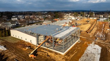 Çalışmalar büyük bir ticari yapı üzerinde devam ederken, işçiler aktif olarak çatı malzemeleri kuruyor ve bulutlu bir gökyüzü altında çelik iskeleti güvence altına alıyorlar. Çevre tarlalar ve evler görülebilir.