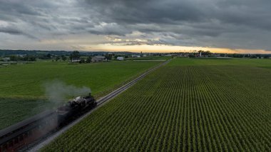 Kaliteli bir buhar treni, canlı yeşil alanlarla çevrili demiryolu rayları boyunca tıkınır. Gökyüzü, arka planda güneş batarken dramatik bulutlarla doludur. Resimli bir kırsal manzara yaratır..