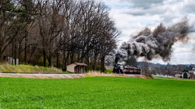 Antika bir buhar treni, canlı yeşil bir arazinin yanındaki raylar boyunca hareket ederken siyah dumanı dalgalandırır. Yakınlarda ağaçlar ve küçük bir barınak kırsal alanı tamamlıyor..