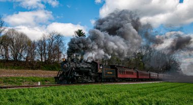Ronks, Pennsylvania, 13 Nisan 2025 - Buharlı bir tren güneşli bir günde pitoresk yeşil bir arazide seyahat ederken siyah duman püskürtür. Ağaçlar, mavi gökyüzünün altında kabarık arkaplan oluşturur.