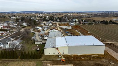 İnşaat işçileri, kırsal bir alanda evler ve tarım arazileriyle çevrili büyük bir depo alanında meşguller..