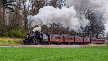 Ronks, Pennsylvania, 5 Nisan 2024 klasik bir buhar treni raylar boyunca tıkırdıyor ve mavi gökyüzüne beyaz duman püskürtüyor. Trenin etrafını çevreleyen yeşil alanlar manzaralı bir zemin oluşturuyor.