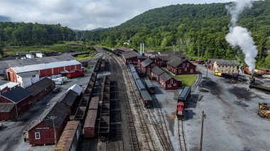 İyi korunan bir demiryolu sahasında kırsal kırmızı binaların yanına park edilmiş vintage trenler yer alıyor. Çevresindeki yemyeşil tepeler buharla dolup taşıyor..