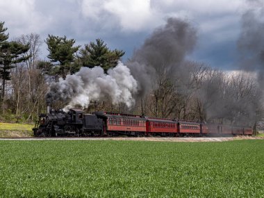 Antika bir buharlı lokomotif, manzaralı bir yol boyunca kırmızı bir yolcu vagonunu çeker. Tren yeşil alan ve bulutlu gökyüzüne duman bulutları yayar ve bir anlığına nostalji yakalar..