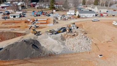 Ağır makineler kırsal bir yerdeki bir inşaat sahasında büyük miktarda ezilmiş taş ve toprak işliyor. İş ilerledikçe kamyonlar malzemeleri hareket ettirir.