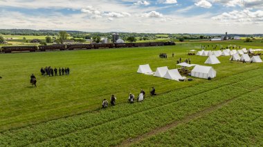 Ronks, Pennsylvania, 9 Haziran 2024 - Dönem kostümleri içindeki katılımcılar, bir İç Savaş canlandırması için yemyeşil bir alanda toplanıyorlar. Çadırlar ve tarihsel gösteriler, iç içe geçen bir atmosfer yaratıyor.