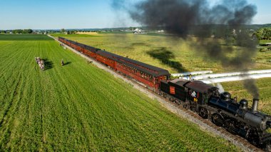 Ronks, Pennsylvania, 9 Haziran 2024 - Bir buhar treni, geniş yeşil alanlarla çevrili manzaralı bir rota boyunca klasik yolcu vagonlarını çeker. İnsanlar bu güzel günde gezintiden ve manzaradan hoşlanırlar.