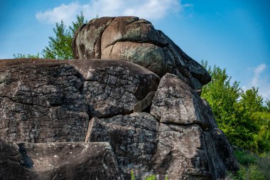 Yüze benzeyen çarpıcı bir kaya oluşumu parlak mavi bir gökyüzünün altında dokulu taş yüzeylerle belirgin bir şekilde sergilenir. Yeşillik bölgeyi sarmış, doğal güzelliği arttırıyor..
