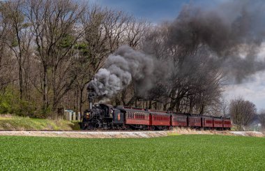 Ronks, Pensilvanya, 4 Nisan 2024 - Eski model bir buhar treni kırsal bir yolda ilerliyor, dalgalanan siyah dumanlar yayıyor. Trenin etrafını saran yemyeşil alanlar bulutlu gökyüzü ve yapraksız yapraklarla kaplı.