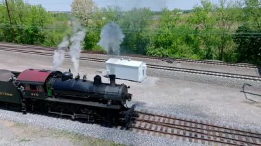 Cennet, Pennsylvania, 17 Mayıs 2023 - Klasik bir buhar lokomotifi, yemyeşil ağaçlarla çevrili demiryolu rayları boyunca ilerliyor. Buhar küçük bir barakanın yanından geçerken havayı dolduruyor..