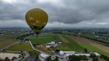 Canlı bir sıcak hava balonu yemyeşil tarlaların ve bulutlu bir gökyüzünün altındaki kırsal binaların üzerinde sürüklenir. Bu sakin manzara sonbaharda kırsal kesimin güzelliğini yansıtıyor..