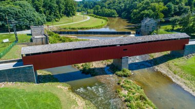 Çarpıcı kırmızı bir köprü, sakin bir nehrin üzerinde, kırsal bir alanda uzanıyor. Bereketli yeşillik, açık güneşli havalarda huzurlu atmosferi artırır..
