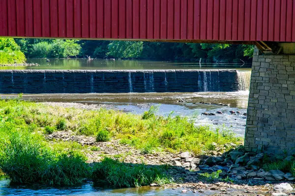 Kırmızı kaplı bir köprü, alçak bir barajın üzerinde çağlayan suyun olduğu sakin bir nehrin üzerinde kemer şeklinde uzanır. Bereketli yeşillik, huzurlu bir kırsal bölgede yazın güzelliğini vurguluyor..