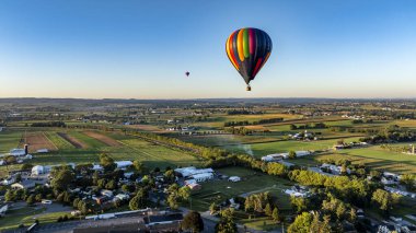 İki canlı sıcak hava balonu, dingin kırsal bölgede zarif bir şekilde yükselerek, açık mavi gökyüzünün altındaki geniş tarlaların ve çiftliklerin güzelliğini sabahın erken saatlerinde yakalıyorlar..
